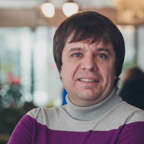 Portrait of Oleksii (Alex) Vinogradov smiling, in a grey and purple sweater, in a softly lit room.