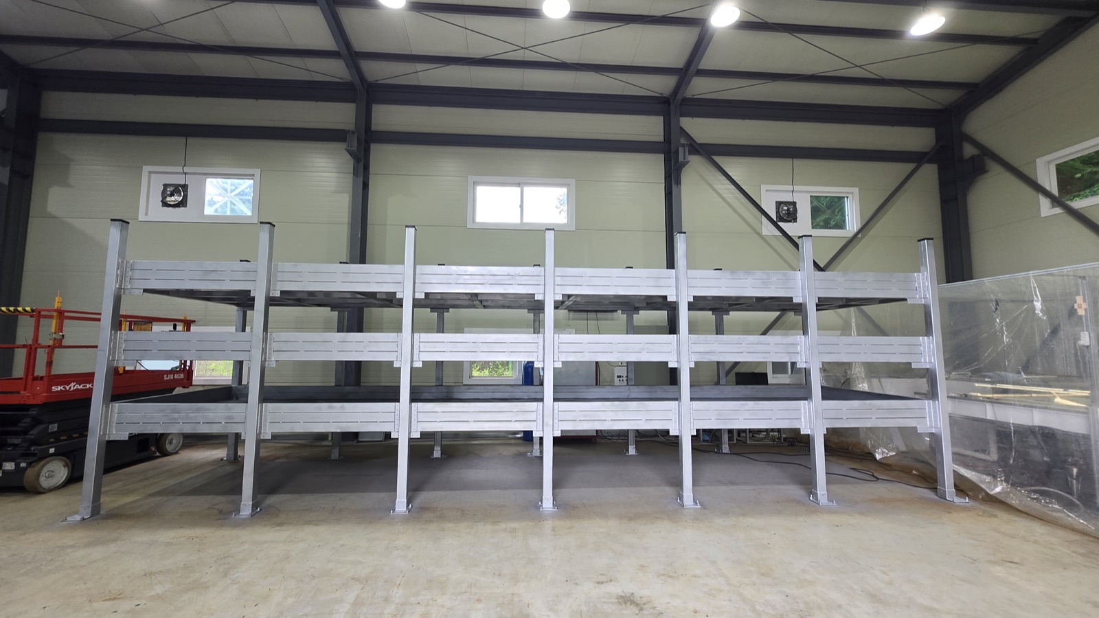 Three rows of empty grey metal racks, stacked three levels high, standing in a large clean warehouse with a steel roof and bright ceiling lights.