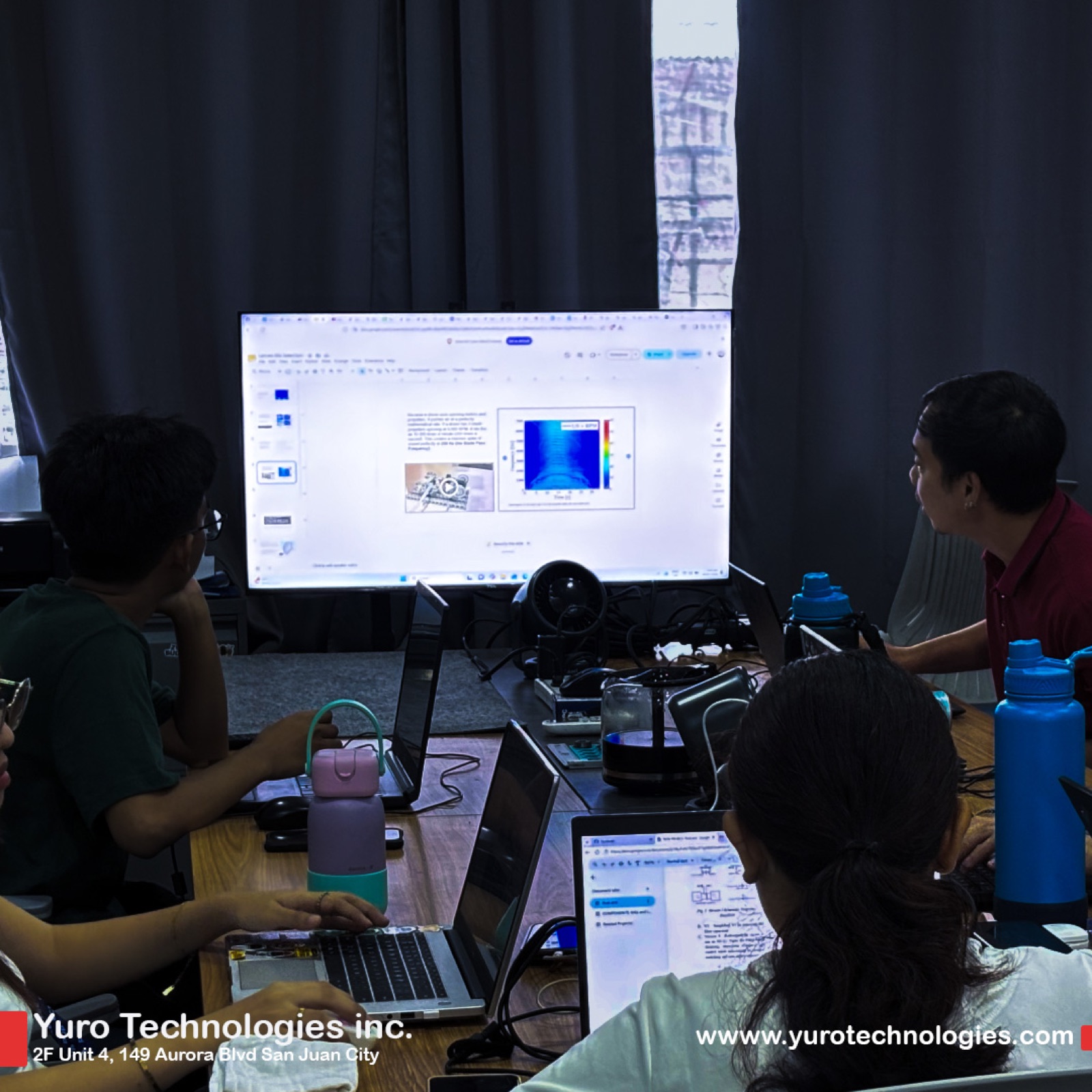 Young people working at laptops around a long table, with a large monitor showing a heat map and small devices on the desk.